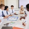 Female High School Teacher Sitting At Table With Teenage Pupils Wearing Uniform Teaching Lesson Female High School Teacher Sitting At Table With Teenage Pupils Wearing Uniform Teaching Lesson