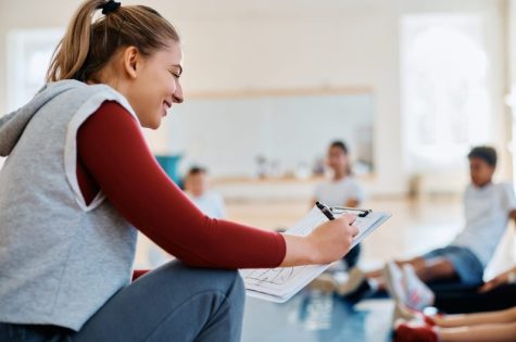 Happy female coach analyzing exercise plan while having physical education class with group of kids at school gym.